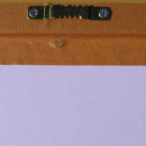 May include: A close-up of a brown wooden frame with a metal hanging hook attached to the back. The frame is covered in a light brown paper tape.