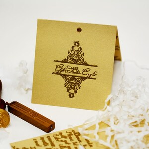 May include: A brown wooden stamp with a gold wax seal and a wooden handle. The stamp is next to a brown paper tag with a decorative design and the text "Gift Studio Craft".