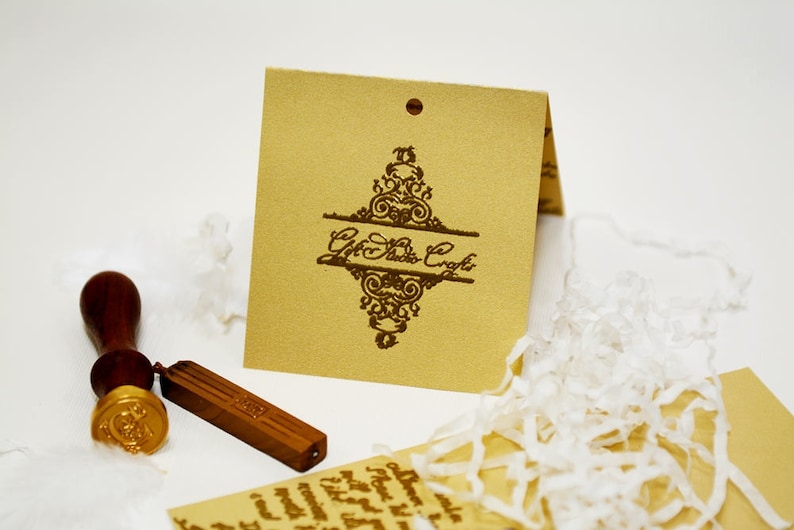 May include: A brown wooden stamp with a gold wax seal and a wooden handle. The stamp is next to a brown paper tag with a decorative design and the text "Gift Studio Craft".