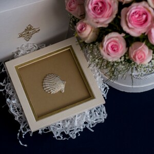 May include: A white picture frame with a gold trim, containing a white seashell on a brown background. The frame is sitting on white shredded paper, next to a white box with pink roses.