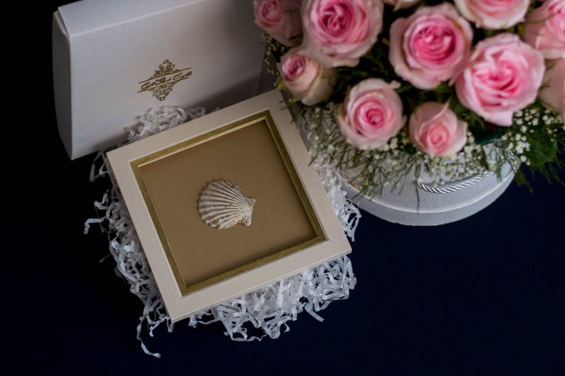 May include: A white picture frame with a gold trim, containing a white seashell on a brown background. The frame is sitting on white shredded paper, next to a white box with pink roses.