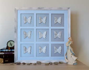 May include: A square wall art piece featuring a distressed white wooden frame enclosing nine individual light blue compartments, each containing a single white butterfly figure. The arrangement is displayed alongside a stack of books and a figurine.