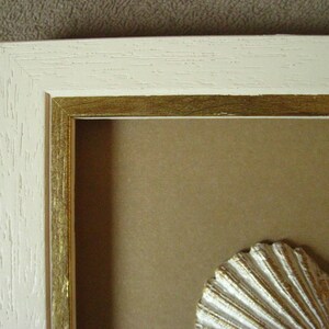 May include: A white picture frame with a gold inner border. The frame is empty, but there is a white seashell in the bottom right corner.