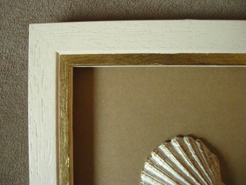 May include: A white picture frame with a gold inner border. The frame is empty, but there is a white seashell in the bottom right corner.