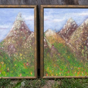 May include: Two framed paintings depicting a mountain landscape with green foliage and a blue sky. The paintings are in gold frames and are displayed against a wooden background.