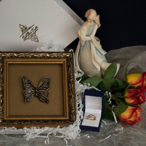 May include: A gold-framed picture with a butterfly inside, a blue ring box with two gold rings, and a bouquet of orange roses. The picture is sitting on a white surface with white tissue paper.