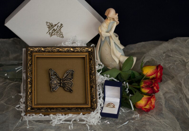 May include: A gold-framed picture with a butterfly inside, a blue ring box with two gold rings, and a bouquet of orange roses. The picture is sitting on a white surface with white tissue paper.