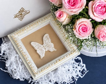 May include: A square white and gold ornate frame containing a white butterfly cutout on a tan background. The frame rests on white shredded paper next to a bouquet of pink roses. A white box with a gold logo is visible in the background.