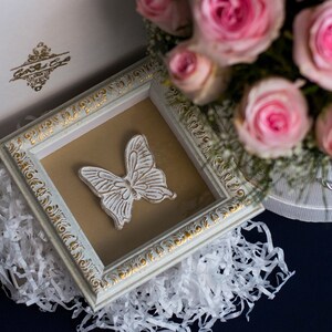 May include: A white framed picture with a white butterfly design on a brown background. The frame has a gold trim and is surrounded by white tissue paper.