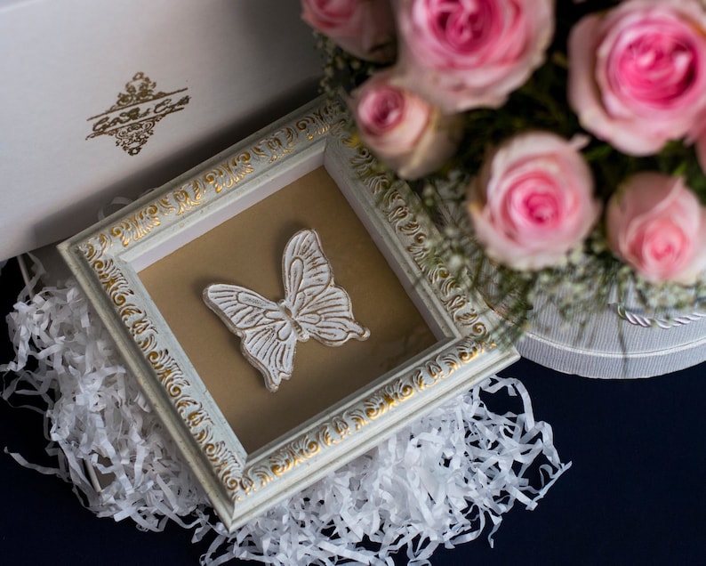 May include: A white framed picture with a white butterfly design on a brown background. The frame has a gold trim and is surrounded by white tissue paper.