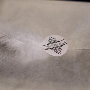 May include: A white feather lies on a white textured surface with a black and white sticker that says "Gift Studio Crafts".
