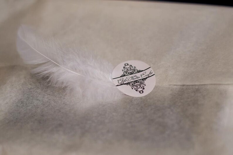 May include: A white feather lies on a white textured surface with a black and white sticker that says "Gift Studio Crafts".