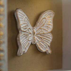 May include: A white butterfly sculpture with gold accents is framed in a white and gold ornate frame.