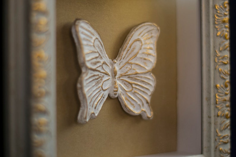 May include: A white butterfly sculpture with gold accents is framed in a white and gold ornate frame.