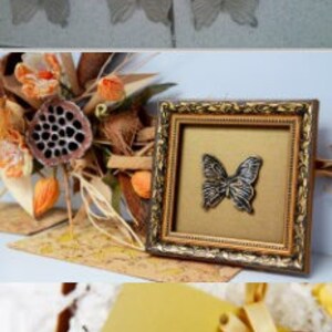May include: A gold-framed picture with a black butterfly design on a gold background. The frame has a decorative, ornate design. The picture is sitting on a table with a gold and white card with a decorative design.