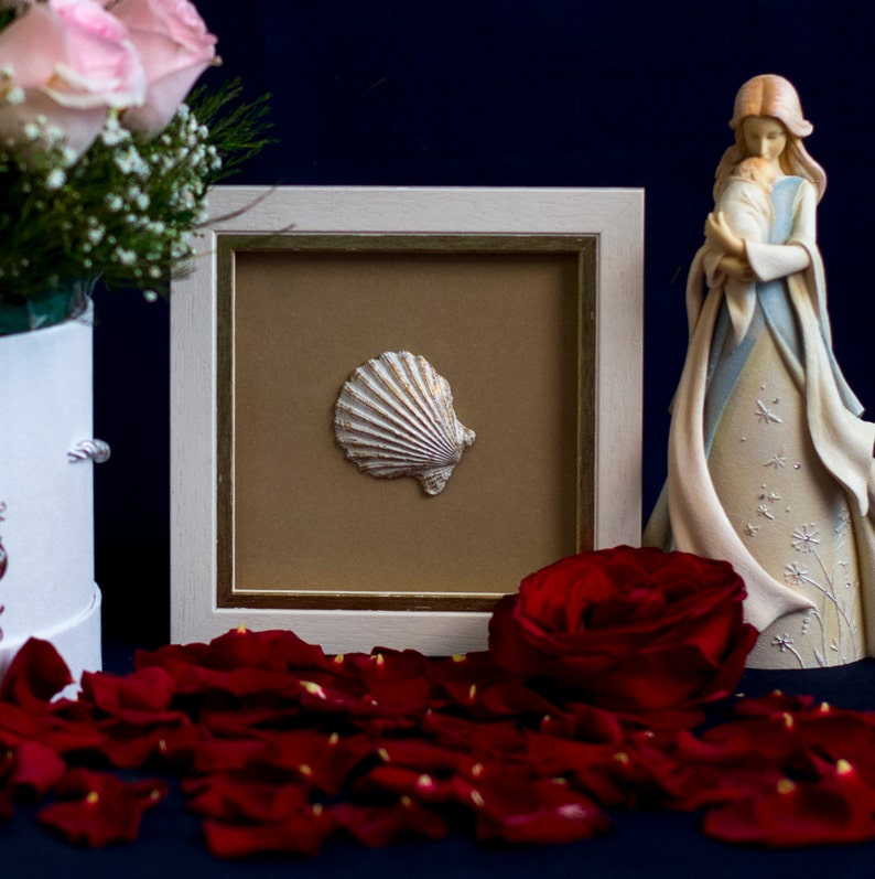 May include: A white seashell framed in a white frame with a gold trim. The frame is sitting on a table with red rose petals scattered around it. A white figurine of a woman holding a baby is in the background.