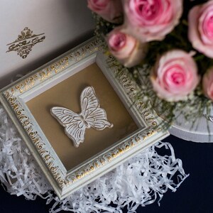 May include: A white framed picture with a white butterfly design on a brown background. The frame has a gold trim and is surrounded by white tissue paper. A bouquet of pink roses is in the background.