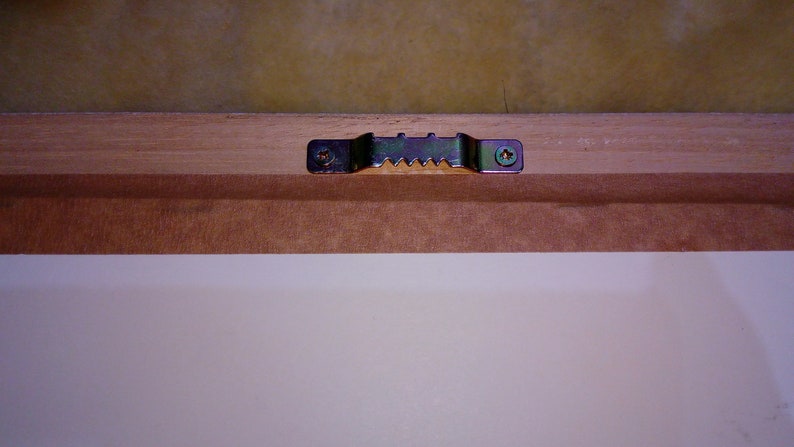 May include: A close-up of a wooden picture frame with a metal hanging hook attached to the back. The hook is secured with two screws.