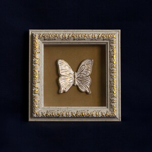 May include: A white butterfly with gold accents is displayed in a white frame with gold trim. The frame has a decorative design.