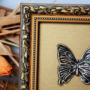 May include: A gold picture frame with ornate black detailing and a black butterfly decoration on the inside.