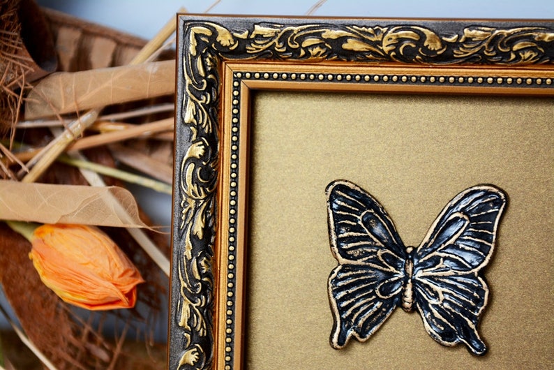 May include: A gold picture frame with ornate black detailing and a black butterfly decoration on the inside.