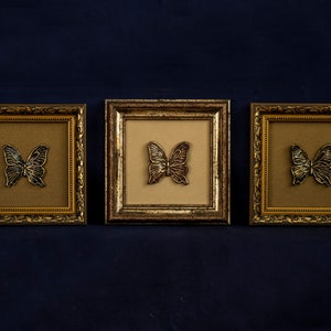 May include: Three gold-colored picture frames with ornate detailing, each featuring a black butterfly silhouette on a beige background.