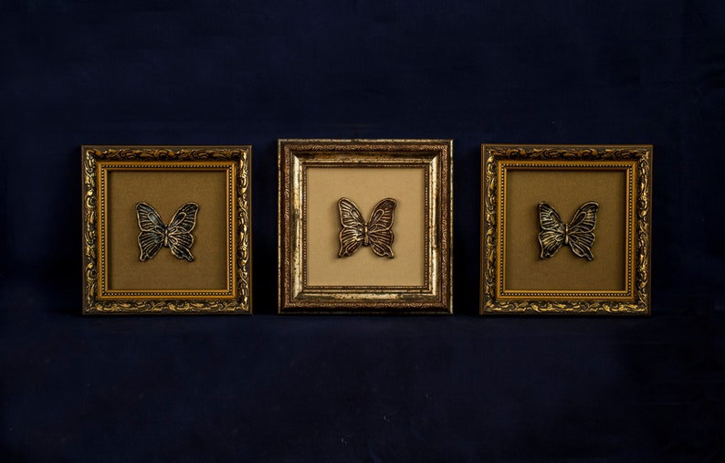 May include: Three gold-colored picture frames with ornate detailing, each featuring a black butterfly silhouette on a beige background.