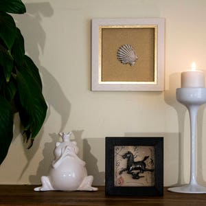 May include: A white ceramic frog figurine with a crown on its head sits on a wooden surface next to a black framed picture with a black and white image of a horse. A white candle holder with a lit candle sits on the right side of the image.