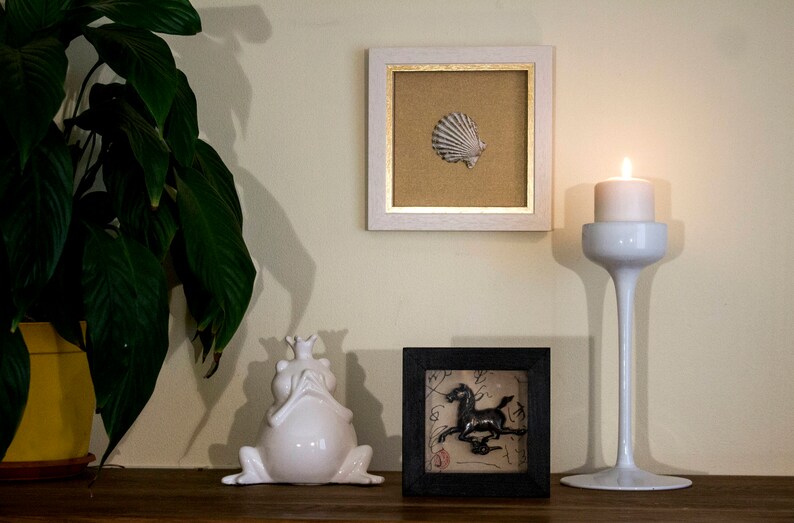May include: A white ceramic frog figurine with a crown on its head sits on a wooden surface next to a black framed picture with a black and white image of a horse. A white candle holder with a lit candle sits on the right side of the image.