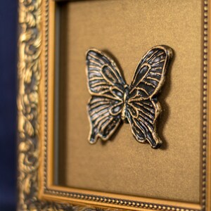 May include: A black butterfly sculpture in a gold ornate frame. The butterfly is raised from the background and has a detailed design.