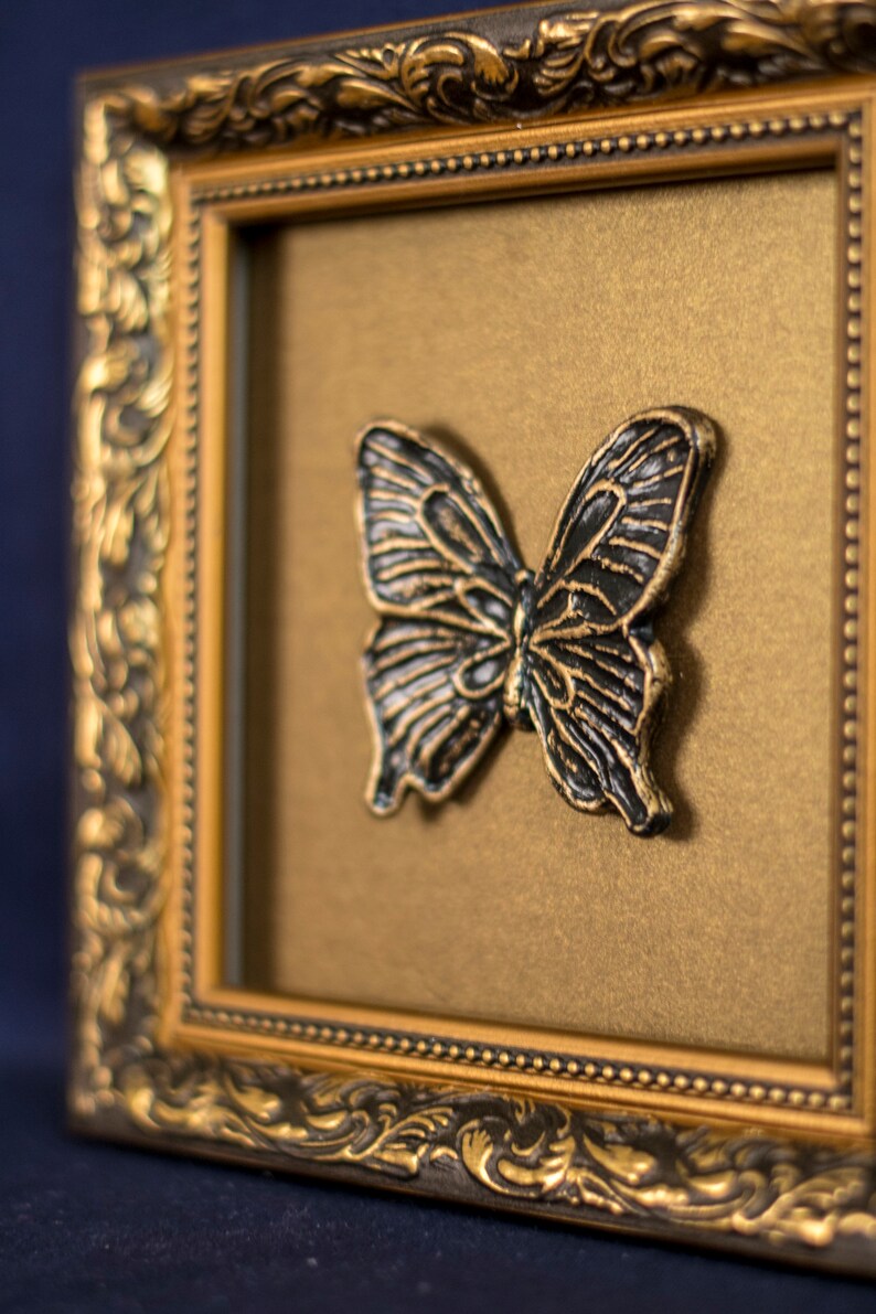 May include: A black butterfly sculpture in a gold ornate frame. The butterfly is raised from the background and has a detailed design.