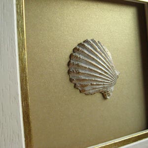 May include: A white picture frame with a gold trim surrounds a gold background with a white seashell in the center.