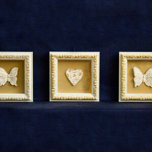 May include: Three white picture frames with gold trim, each featuring a different white decorative element: a butterfly, a heart, and a butterfly. The frames are displayed against a dark blue background.