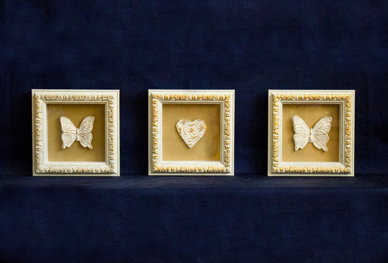 May include: Three white picture frames with gold trim, each featuring a different white decorative element: a butterfly, a heart, and a butterfly. The frames are displayed against a dark blue background.