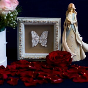 May include: A white framed picture with a white butterfly design, a red rose, and a figurine of a woman holding a baby. The figurine is standing on a bed of red rose petals.