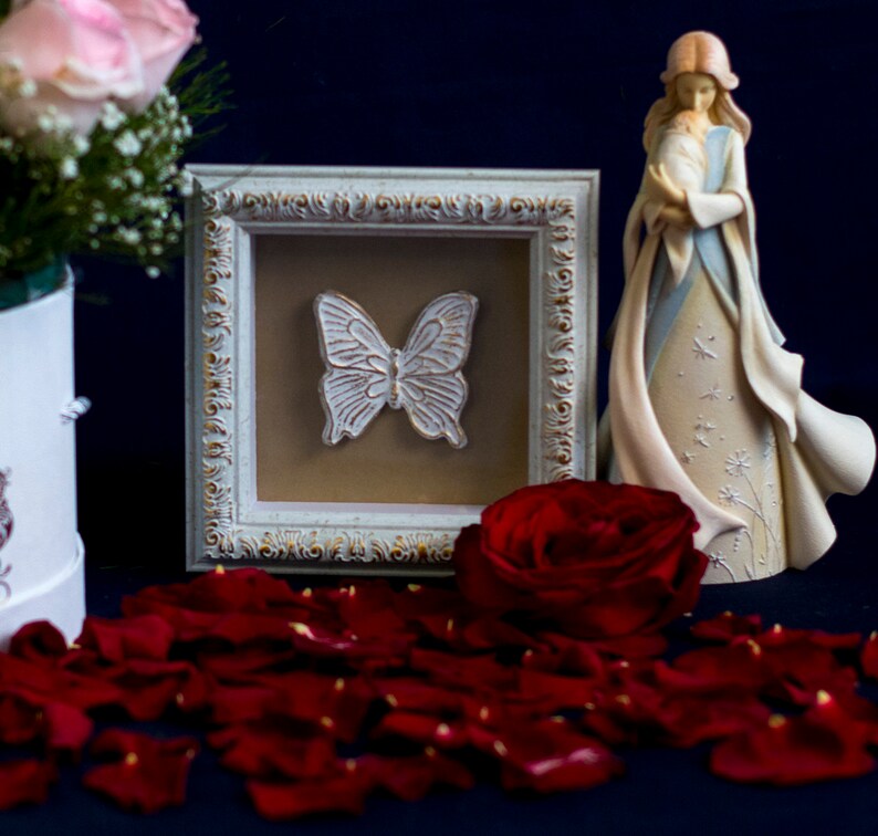 May include: A white framed picture with a white butterfly design, a red rose, and a figurine of a woman holding a baby. The figurine is standing on a bed of red rose petals.