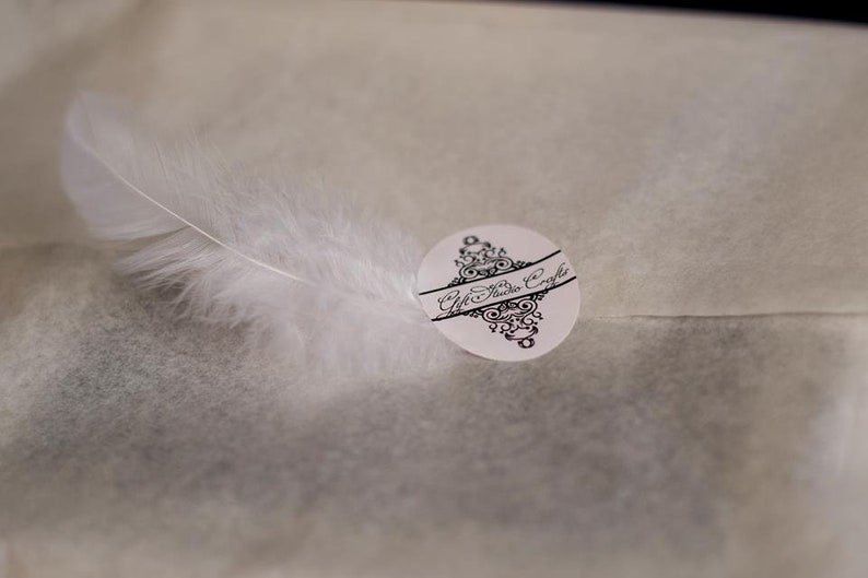 May include: A white feather sits on a white tissue paper background with a black and white sticker that says "Gift Studio Crafts".