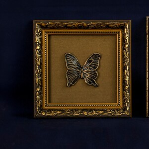 May include: A gold-colored ornate picture frame with a black butterfly design inside. The frame has a brown mat and a gold-colored border.