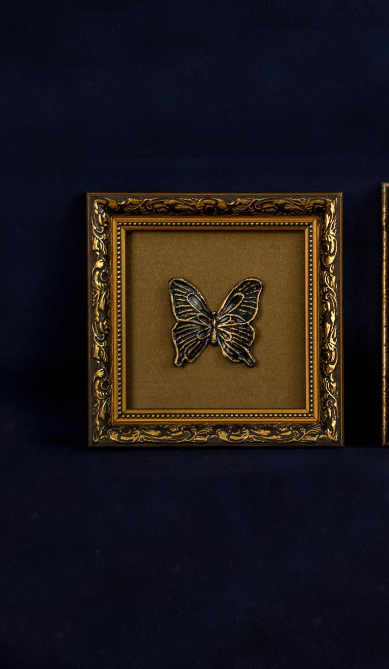 May include: A gold-colored ornate picture frame with a black butterfly design inside. The frame has a brown mat and a gold-colored border.