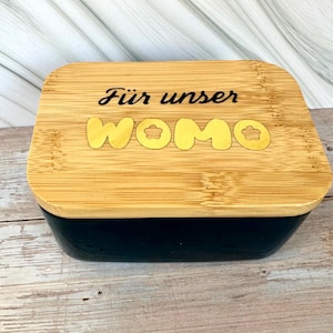 Könnte beinhalten: Eine rechteckige schwarze Bambusbox mit einem Bambusdeckel. Der Deckel trägt die Worte "Für unser WOMO" in schwarzen und gelben Buchstaben. Die Box steht auf einer hellbraunen Holzoberfläche.