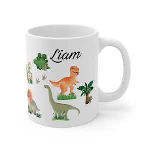 Könnte beinhalten: Weißer Keramikbecher mit bunten Cartoon-Dinosauriern und dem Namen "Liam" in schwarzer Schrift. Die Dinosaurier sind in verschiedenen Farben, darunter Grün und Orange. Der Becher hat einen weißen Henkel.