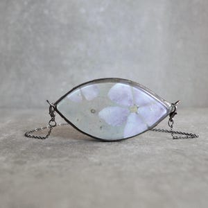 Puede incluir: Un colgante en forma de marquesa que presenta una flor prensada de color morado claro incrustada en resina transparente con un borde metálico oscuro. El colgante está unido a una fina cadena metálica oscura a ambos lados, creando un collar horizontal.