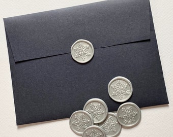 May include: A black envelope with silver snowflake wax seals. The envelope is closed and the seals are scattered around it.