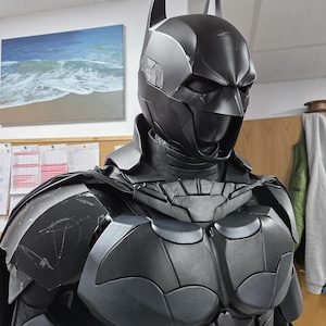 May include: A black Batman costume with a cowl featuring pointed ears. The armor-like chest piece has a bat symbol. The costume is dark gray and black, with a textured appearance. The image is a close-up of the costume.