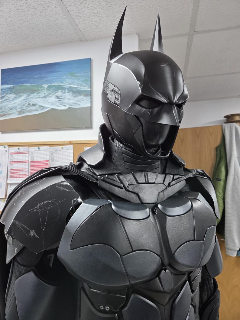May include: A black Batman costume with a cowl featuring pointed ears. The armor-like chest piece has a bat symbol. The costume is dark gray and black, with a textured appearance. The image is a close-up of the costume.