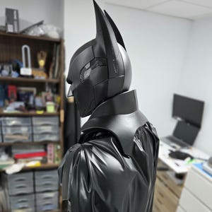 May include: A black Batman cowl and cape set. The cowl features pointed ears and a segmented design with a textured pattern on the side. The cape is made of a shiny, flowing material, creating a dramatic silhouette.