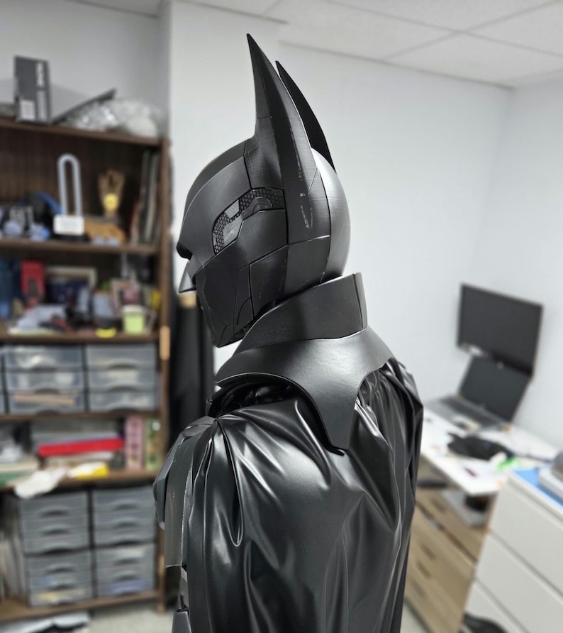 May include: A black Batman cowl and cape set. The cowl features pointed ears and a segmented design with a textured pattern on the side. The cape is made of a shiny, flowing material, creating a dramatic silhouette.