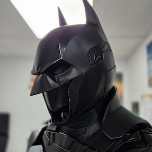 May include: A black Batman cowl and chest armor set. The cowl features pointed ears and a textured surface. The chest armor has a segmented design. The overall aesthetic is dark and imposing, with a focus on detail.
