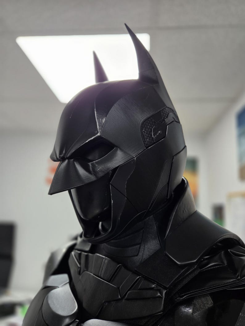 May include: A black Batman cowl and chest armor set. The cowl features pointed ears and a textured surface. The chest armor has a segmented design. The overall aesthetic is dark and imposing, with a focus on detail.