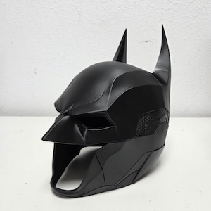 May include: A black Batman cowl, featuring a pointed nose, eye openings, and tall, pointed ears. The mask has a textured surface and is designed to fit the head. The overall design is sleek and angular.